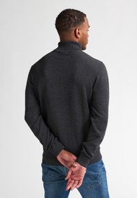 Suéter gris oscuro con cuello alto, mangas largas y puños acanalados. Llevado con jeans azules, mostrando una textura lisa y sencilla.