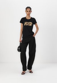 T-shirt nera con stampa grafica dorata, abbinata a pantaloni neri a vita alta. La modella tiene una borsa nera e indossa tacchi neri.