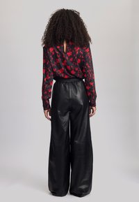 Schwarze Bluse mit floralen Motiven und roten Mustern, kombiniert mit hochgeschnittenen, weiten schwarzen Lederhosen. Schlüssel-Ausschnitt am Kragen.