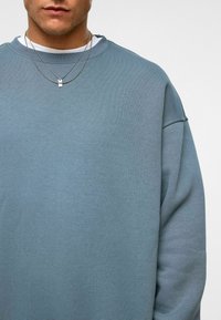 Lichtblauwe katoenen sweatshirt met een relaxte pasvorm, geribbelde ronde hals en verlaagde schouders. Gelaagd met twee zilveren kettingen.