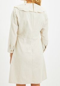 Vestido beige claro con mangas largas y un cuello de encaje con bordes en forma de ola, que presenta un cierre en la parte posterior y puños abotonados, hecho de una tela texturizada.