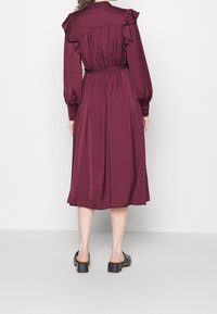 Burgundské šaty s dlouhými rukávy, volánkovými rameny a páskem v pase. Vyrobené z hladkého materiálu, sahající po lýtka. Černé sandály na silném podpatku.