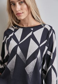 Himmelblaues und hellgraues Langarmshirt mit einem geometrischen Muster aus unterschiedlichen dreieckigen Formen und Texturen über den Stoff verteilt.