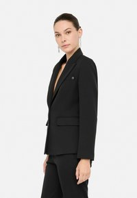 Černý vypasovaný blazer s klopovým límcem, zapínání na jeden knoflík, dvěma předními kapsami a hladkou, strukturovanou texturou látky.