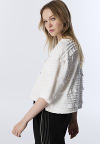 Top bianco e testurizzato con volant orizzontali, scollatura rotonda e maniche a tre quarti, abbinato a pantaloni neri con strisce bianche sui lati.