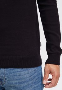 Suéter de punto negro con dobladillo y puños de canalé, que presenta una textura sutil. En el lateral tiene una pequeña etiqueta con un patrón de cuadros.