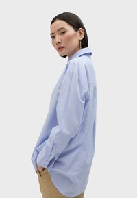 Camicia a righe azzurro chiaro oversized con colletto a camicia e maniche lunghe, caratterizzata da sottili dettagli ricamati e un orlo curvo.