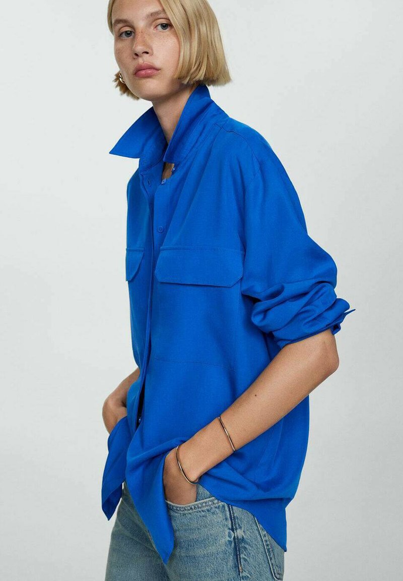 Camicia blu a maniche lunghe con colletto strutturato, due tasche frontali e maniche arrotolate, abbinata a jeans in denim blu chiaro.