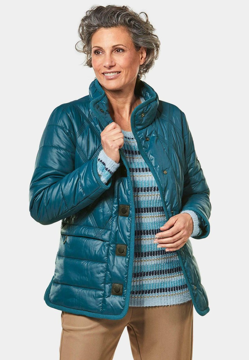 Türkisfarbener gesteppter Mantel mit hohem Kragen, der über einem gestreiften blauen Pullover getragen wird, kombiniert mit beigen Hosen.