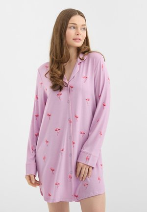 Ung kvinde med langt brunt hår iført en lavendel natkjole med pink flamingomønstre, stående og kiggende til siden på en hvid baggrund.
