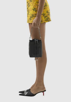 Persoon die een gele minirok met patroon draagt, zwarte muiltjes met puntige hakken en een kleine zwarte handtas met een kettingriem vasthoudt.