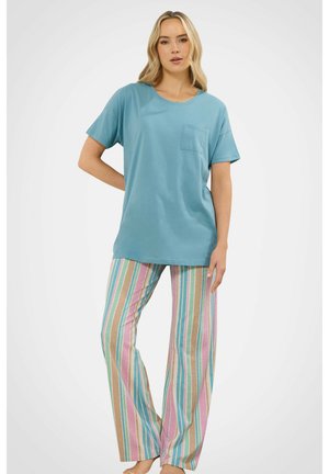 Donna in piedi con la mano sul fianco, indossa una maglietta blu a maniche corte e pantaloni larghi con strisce verticali rosa, blu e beige.