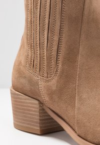 Bottine en suede beige avec des détails de couture verticale, un corps texturé et un petit talon empilé présentant des rainures horizontales.