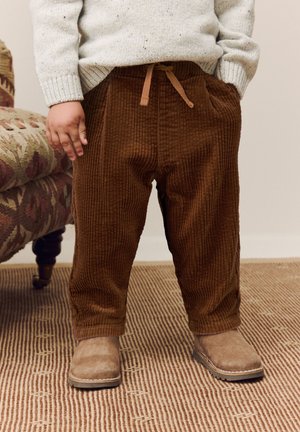 Pantalon en velours côtelé marron avec taille à cordon, jambes larges et poches latérales, associé à des bottes en daim couleur camel. Fond texturé.