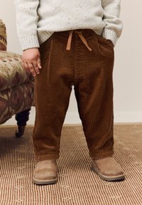 Hnedé kordové nohavice so sťahovacím pásom, s širokými nohavicami a bočnými vreckami, spojené s hnedými semišovými topánkami. Textúrované pozadie.