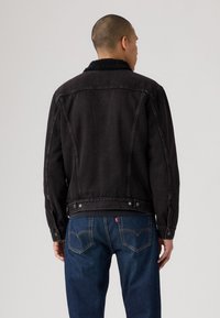 Zwarte spijkerjack met een gestructureerde, harige kraag, zichtbare stiksels en knoopdetails, gedragen met blauwe jeans.