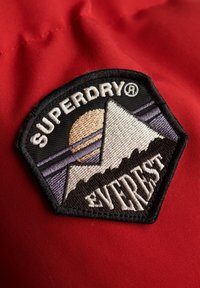 Sechseckiger bestickter Aufnäher auf rotem Stoff mit Berg, Sonne und Text "SUPERDRY® EVEREST" in weißem und schwarzem Faden.
