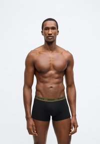 Čierne boxerky s zeleným pásom zdobeným logom "HUGO". Tkanina vyzerá hladko, priliehavý strih, minimálne detaily, dvojfarebný kontrast.