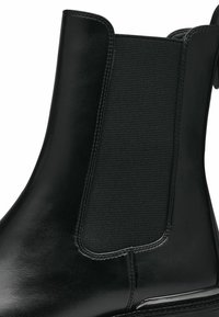 Bota Chelsea negra con un exterior de cuero suave y panel lateral elástico. Presenta una silueta elegante y un acento pulido en la suela.