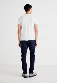 Chemise blanche à manches courtes avec une couture arrière droite, associée à un jean bleu foncé et des chaussures grises, vue de dos.