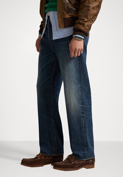 Temno modri jeans iz debelega tekstila s ravno krojem, v kombinaciji z rjavim usnjenim jopičem in rjavimi čevlji loafer. Opažene teksture in detajli šivanja.