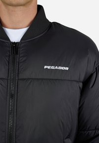 Melnas pūkainā jaka ar ribotu apkakli, priekšējo rāvējslēdzēju un vārdu "PEGADOR", kas izšūts baltā krāsā uz krūškurvja. Tekstūra izskatās gluda un spīdīga.