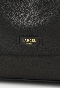 Sac en cuir noir avec une surface texturée, arborant une étiquette de logo doré avec l'inscription "LANCEL PARIS". Détails de couture le long du bord.