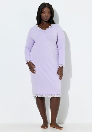 Vrouw staat op blote voeten, gekleed in een lavendelkleurige nachthemd met lange mouwen en een witte kanten afwerking op de kraag, manchetten en zoom, tegen een witte achtergrond.