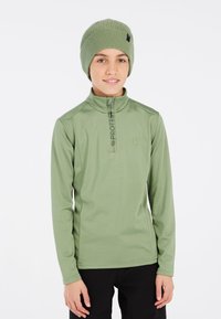 Camisetta a maniche lunghe con zip di colore verde chiaro, realizzata in un tessuto morbido, con colletto alto e dettaglio del logo. Un berretto nero completa l'outfit.