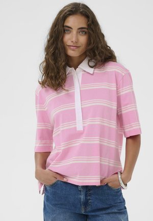 Femme aux cheveux bruns ondulés portant un polo rayé rose et blanc, un jean bleu et un bracelet argenté, les mains dans les poches, souriant doucement.