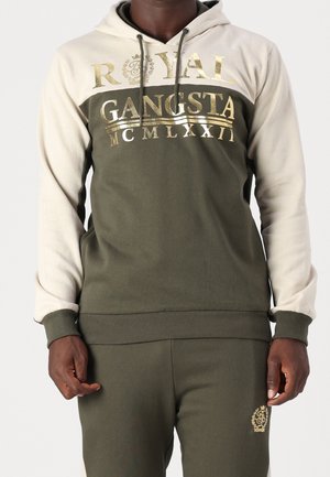 Mężczyzna w oliwkowo-zielonej i beżowej bluzie z kapturem z napisem „ROYAL GANGSTA MCMLXXII” w kolorze złotym oraz dopasowanych oliwkowo-zielonych spodniach z logo na lewej udzie.
