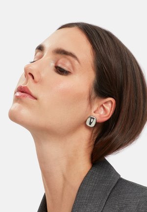 Pendientes cuadrados en blanco y negro con un acabado brillante, con un patrón gráfico único. Llevados por una modelo con cabello corto castaño.