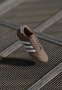 Brown athletic shoe made of suede with white stripes, a textured front, and a smooth heel. Features a laced design and rubber sole.
