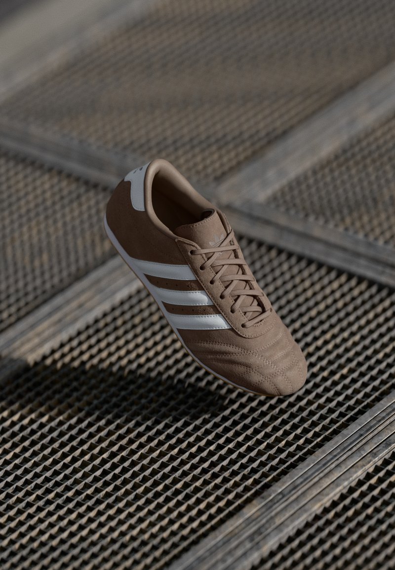 Brown athletic shoe made of suede with white stripes, a textured front, and a smooth heel. Features a laced design and rubber sole.