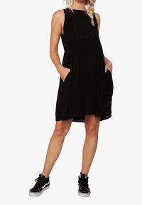 Vestido negro sin mangas con un cuerpo texturizado, cintura caída y bolsillos laterales. Llevado con zapatillas altas negras con suelas blancas.