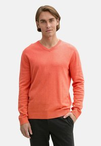 Pulover coral cu decolteu în V, fabricat dintr-un material moale, cu mâneci lungi și manșete și tiv riglate. Modelul poartă pantaloni închiși la culoare, cu mâinile în buzunare.
