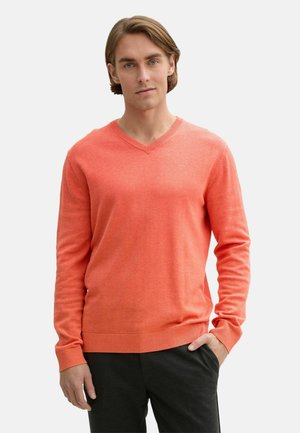 Pulover coral cu decolteu în V, fabricat dintr-un material moale, cu mâneci lungi și manșete și tiv riglate. Modelul poartă pantaloni închiși la culoare, cu mâinile în buzunare.
