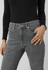 Jeans en denim gris taille haute avec des surpiqûres contrastées, cinq poches et une fermeture à bouton classique, portés avec un haut noir à manches longues.