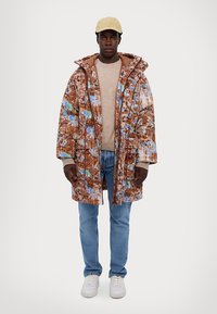 Barna, mintás puff jacket kék és zöld részletekkel, cipzáros elejével és nagy kapucnijával, bézs pulóver és világoskék farmer felett viselve.