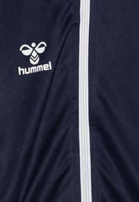 Veste de sport bleu marine en tissu texturé avec une fermeture éclair blanche et le logo Hummel arborant un emblème d'abeille stylisé.