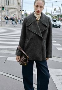 Abrigo de lana gris con diseño de doble botonadura, grandes solapas, combinado con pantalones vaqueros de pierna ancha en denim oscuro y un bolso cruzado marrón.