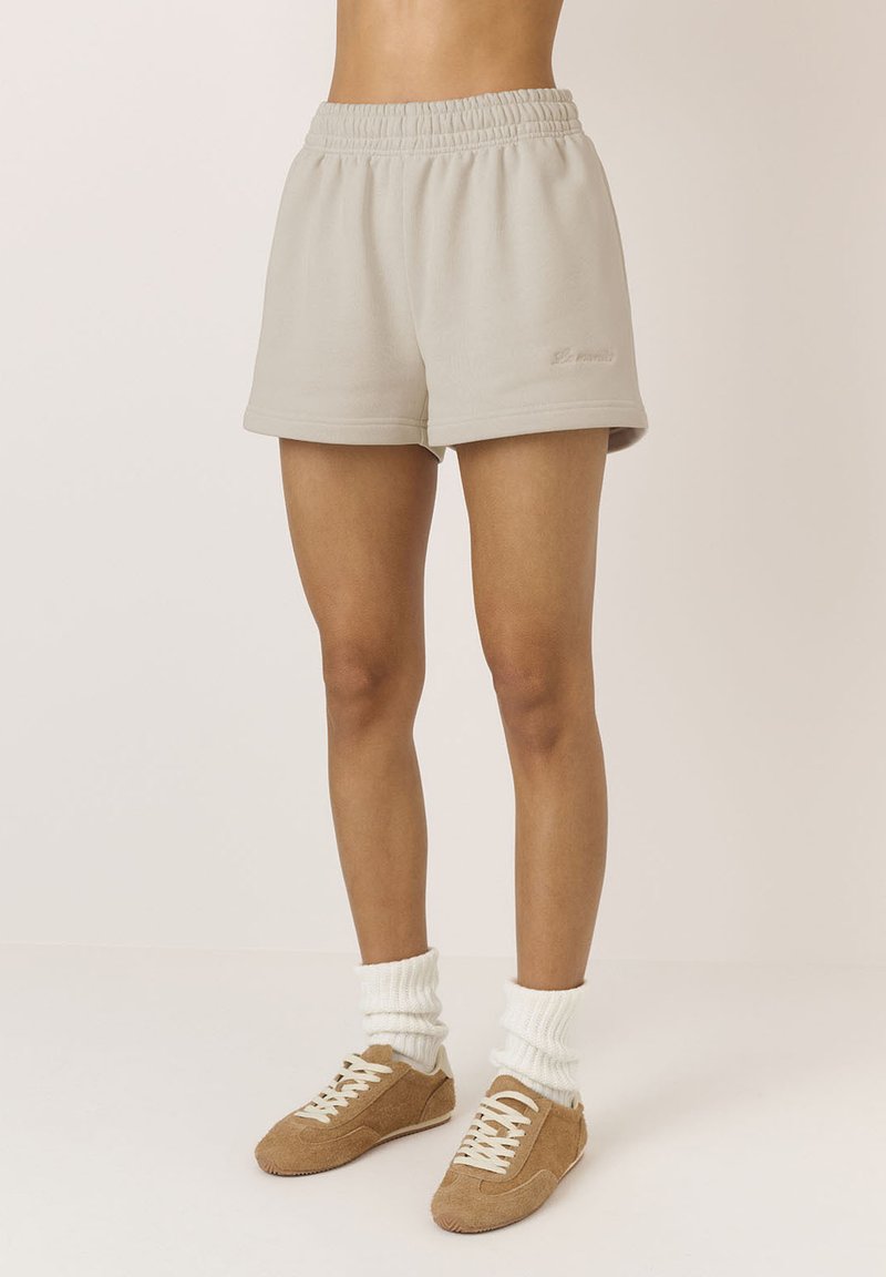 Beżowe bawełniane szorty z elastycznym paskiem w talii i subtelnym logo z przodu. Połączone z puszystymi białymi skarpetkami i brązowymi butami na sznurowadła.