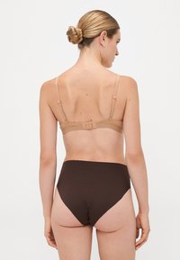 Vrouw met haar in een knot, gekleed in een huidskleurige beha en hoogopgetrokken donkerbruine onderbroek, die met de rug naar de camera staat tegen een effen lichte achtergrond.