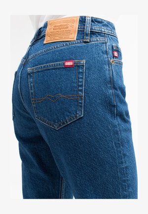 Tumedat sinised teksad tekstuuriga pinnaga, millel on pruun nahklabel, punane silt ja eristuvad õmmeldud tagataskud.