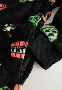 Mustaa fleece-kangasta, jossa on värikkäitä sarjakuvamaisia kuvastoja, mukaan lukien hahmoja ja kuvioita. Näkynä ribatulta mustalta hihalta reunassa lisäyksinä.