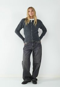 Grau gestrickte Strickjacke mit Knopfverschluss, kombiniert mit lockeren, dunklen Jeans. Das Model steht vor einem weißen Hintergrund.