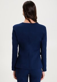 Žena s dlouhými tmavými vlasy, oblečená v obtaženém námořnickém modrém saku a odpovídajících kalhotách, stojí zády k fotoaparátu na jednobarevném pozadí.