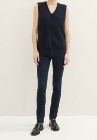 Laivastonsininen neulottu hihaton yläosa, jossa V-kaula-aukko ja ribbineulos, yhdistetty tummiin denimfarkkuihin ja mustiin nauhallisiin kenkiin. Yksivärinen tausta.