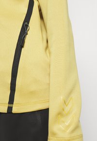 Gelber Jackenärmel mit schwarzem vertikalem Reißverschluss und gelben Chevron-Designs am Handgelenk, getragen über schwarzen Hosen.