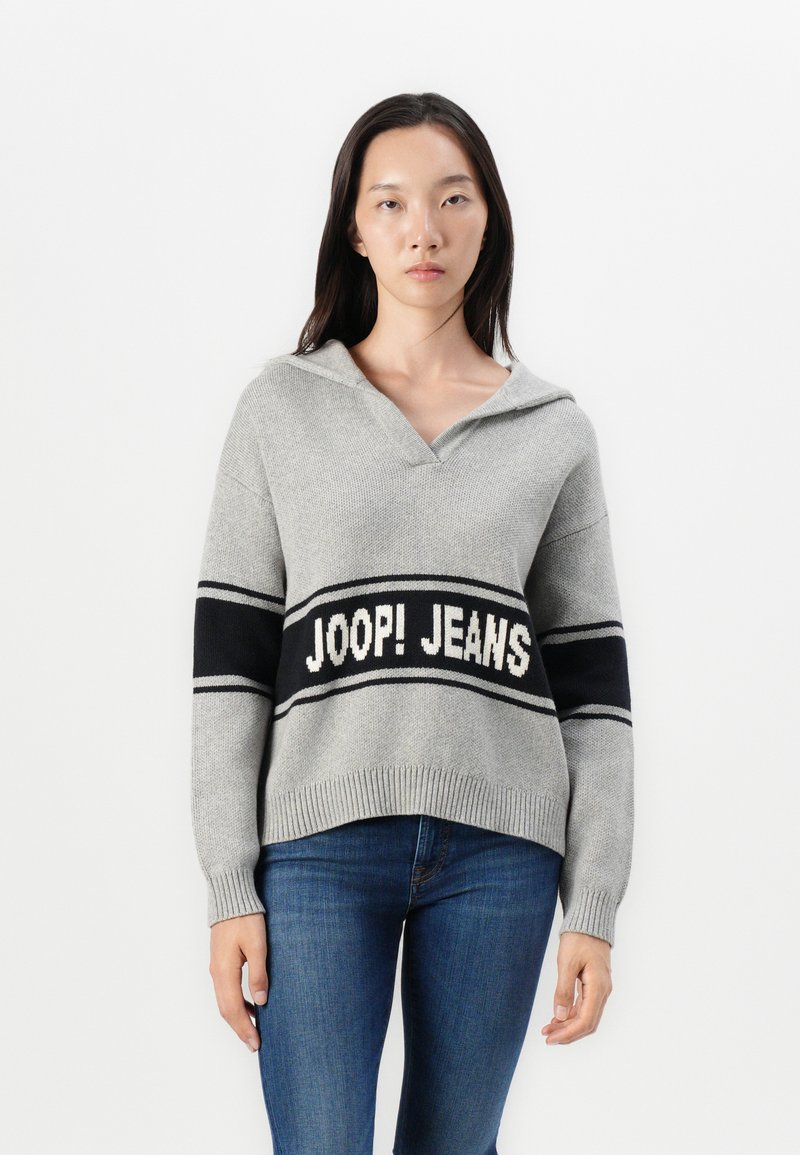 Femme portant un pull en maille gris avec des rayures noires et le texte "JOOP! JEANS", associé à un jean bleu, se tenant contre un fond blanc.